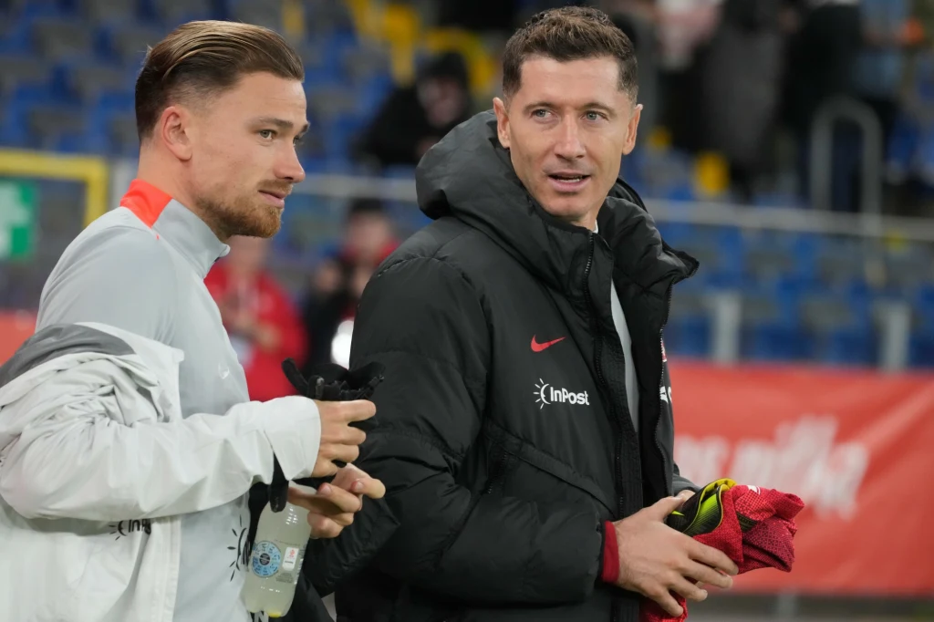 Robert Lewandowski i Matty Cash przed meczem reprezentacji Polski Dwóch mężczyzn stojących na stadionie, ubranych w sportowe kurtki reprezentacyjne, jeden z nich trzyma strój sportowy, drugi rozmawia i trzyma butelkę napoju.