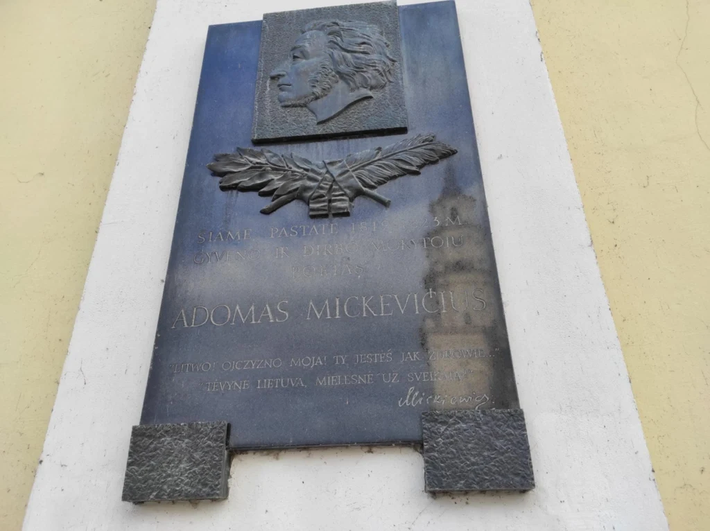 Tablica pamiątkowa z reliefem przedstawiającym profil mężczyzny oraz motyw liścia laurowego, inskrypcja upamiętniająca osobę Adomasa Mickevičiusa, zamontowana na jasnym murze.