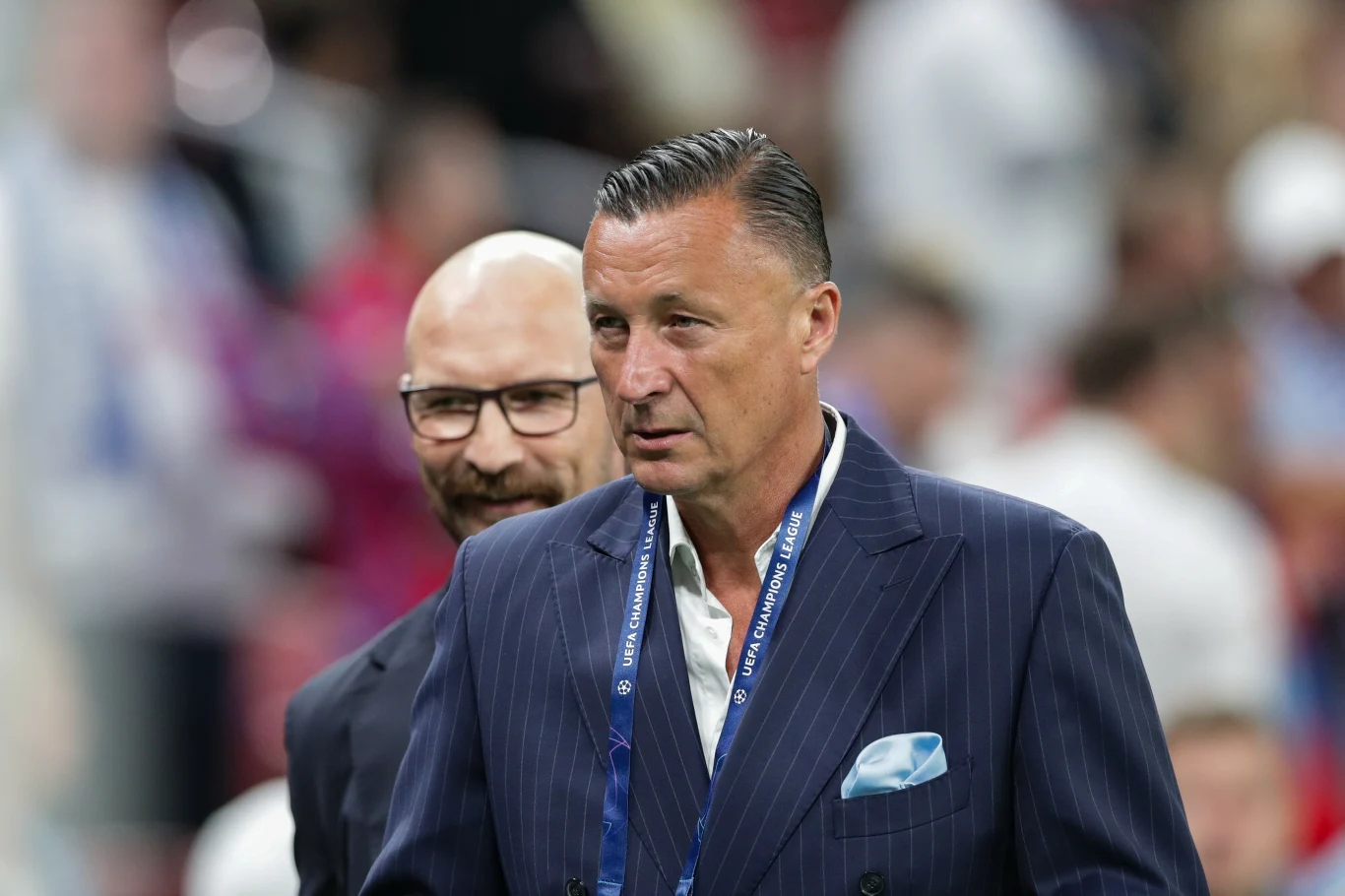 Dwóch elegancko ubranych mężczyzn na tle rozmytego stadionu, jeden w garniturze w prążki z niebieską chusteczką w kieszonce, drugi łysy z brodą w okularach uśmiecha się lekko zza jego pleców.