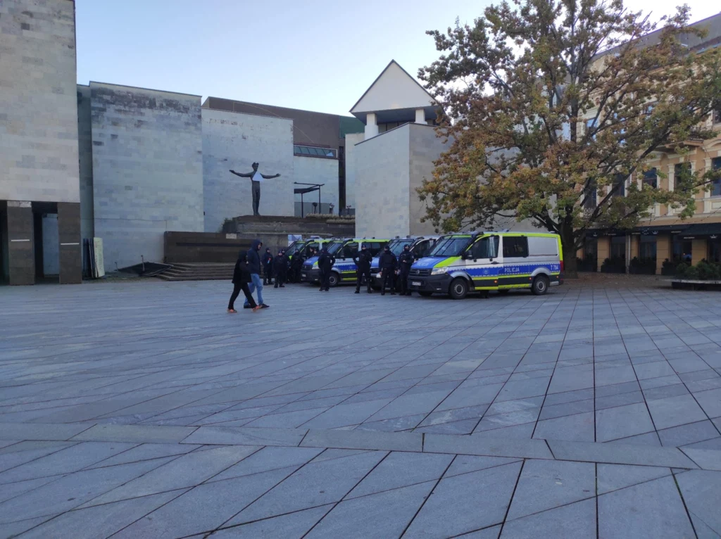 Radiowozy policyjne oraz grupa funkcjonariuszy policji zgromadzeni na placu przed nowoczesnym budynkiem z dużą rzeźbą w tle, jesienno-zielone drzewo po prawej stronie oraz kilka osób przechodzących w pobliżu.