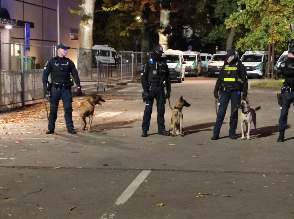 Czterech policjantów z psami służbowymi stojących w rzędzie na ulicy, w tle zaparkowane policyjne pojazdy oraz drzewa oświetlone światłem ulicznym