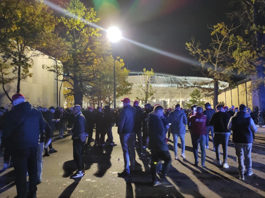 Tłum ludzi zgromadzony na placu podświetlonym światłem ulicznej latarni, część osób ubrana w ciemne kurtki i czapki, w tle widoczny nowoczesny stadion otoczony drzewami.