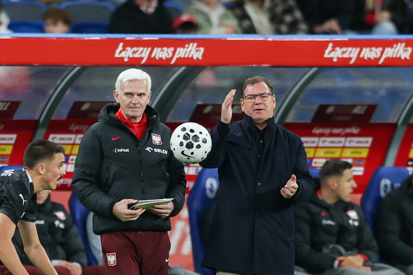 Jan Urban i Jacek Magiera Dwóch mężczyzn stojących przy linii bocznej boiska, z których jeden trzyma tablet ubrany w kurtkę z emblematem reprezentacji Polski, a drugi gestykuluje podczas meczu; w tle ławka rezerwowych oraz napis 'Łączy nas piłka'.