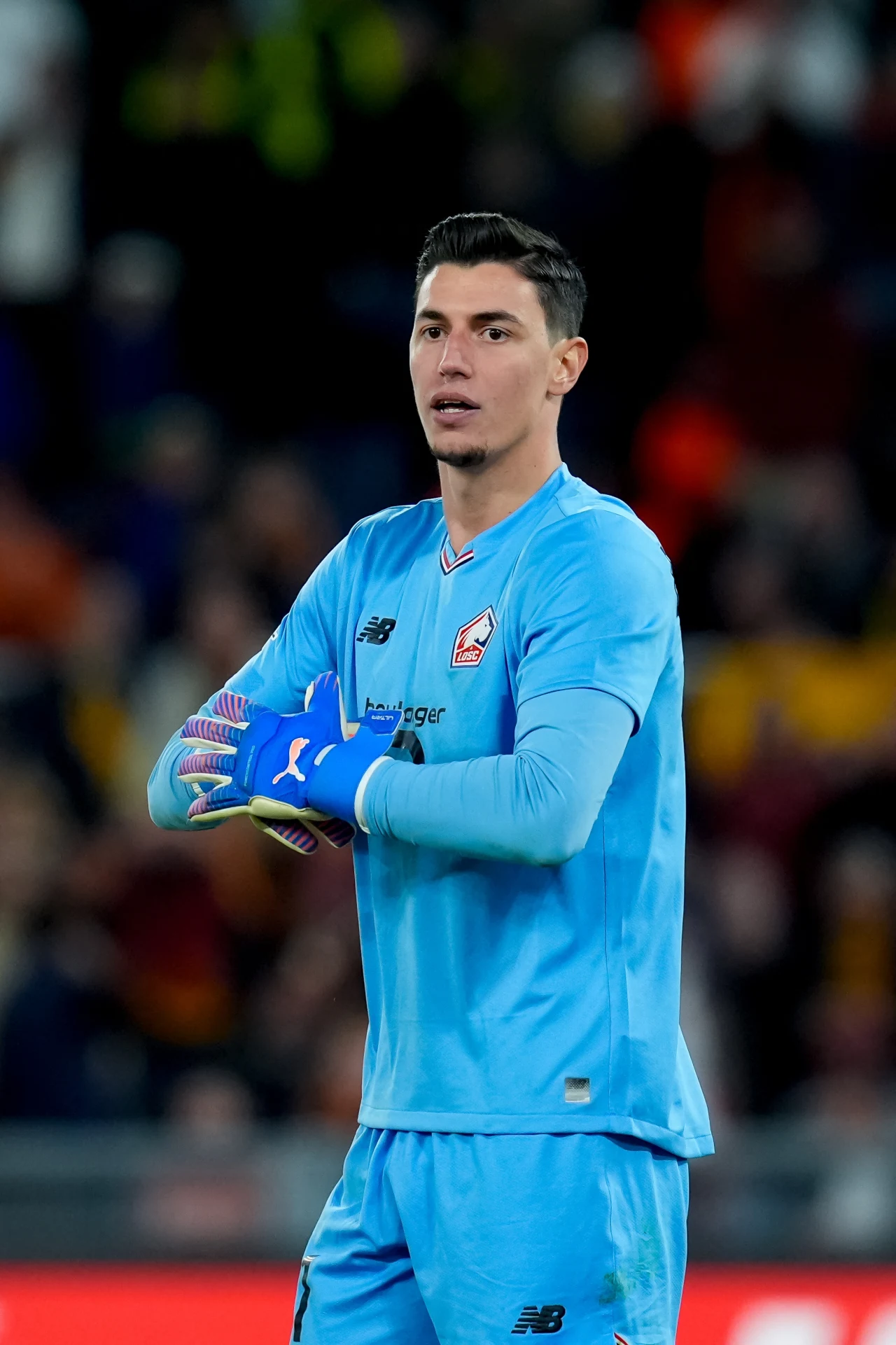 Berke Ozer Bramkarz w niebieskim stroju sportowym stoi na boisku z rękami skrzyżowanymi, nosi niebieskie rękawice bramkarskie, w tle zamazane sylwetki kibiców na trybunach.