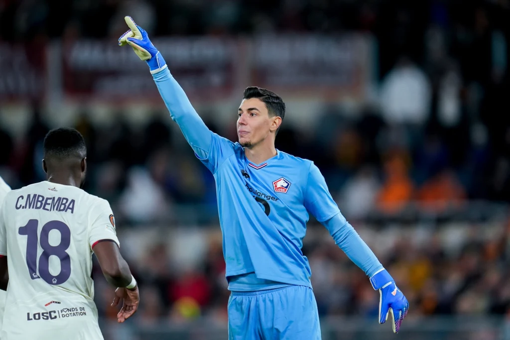 Berke Ozer Bramkarz w niebieskim stroju wydaje polecenia kolegom z drużyny podczas meczu piłkarskiego, obok znajduje się piłkarz w białej koszulce z numerem 18.