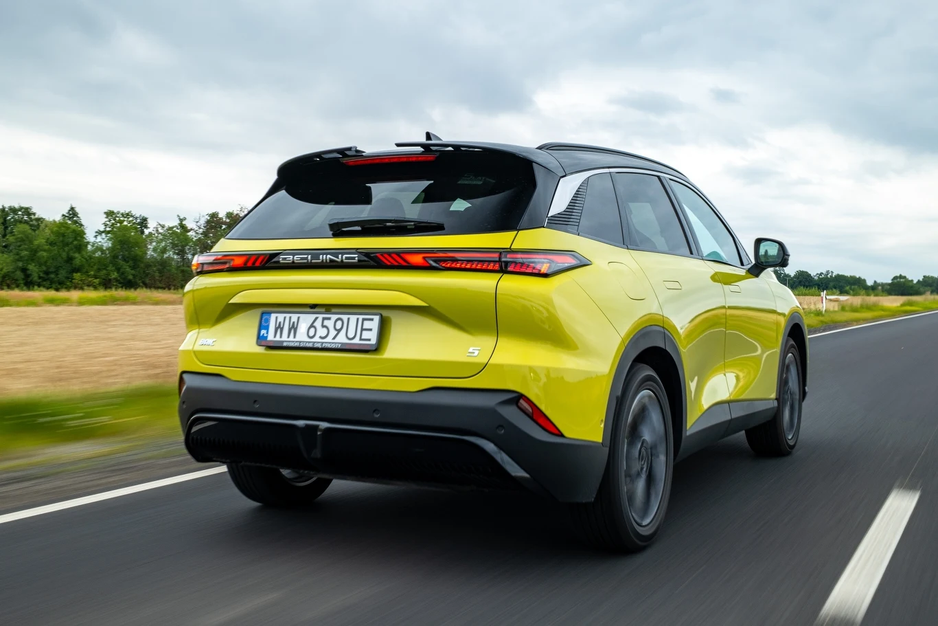 Jasnozielony nowoczesny SUV marki Geely jadący asfaltem przez wiejską okolicę, widok z tyłu z widoczną rejestracją oraz dynamicznym ujęciem samochodu w ruchu