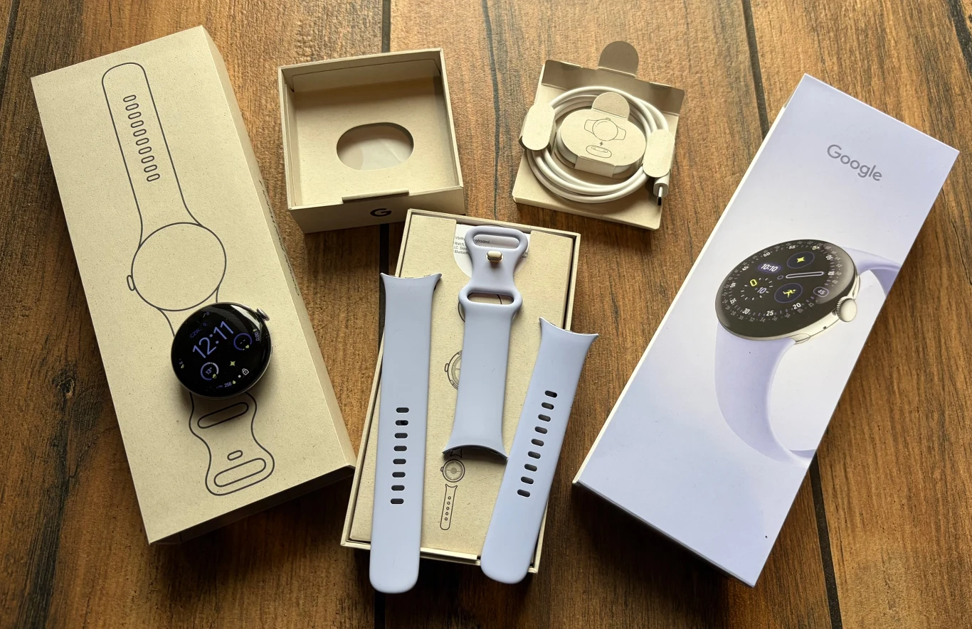 Zawartość opakowania z Google Pixel Watch 4. Opakowanie i zawartość zestawu smartwatcha Google wraz z paskami, stacją ładowania i akcesoriami, wszystko rozłożone na drewnianym stole.