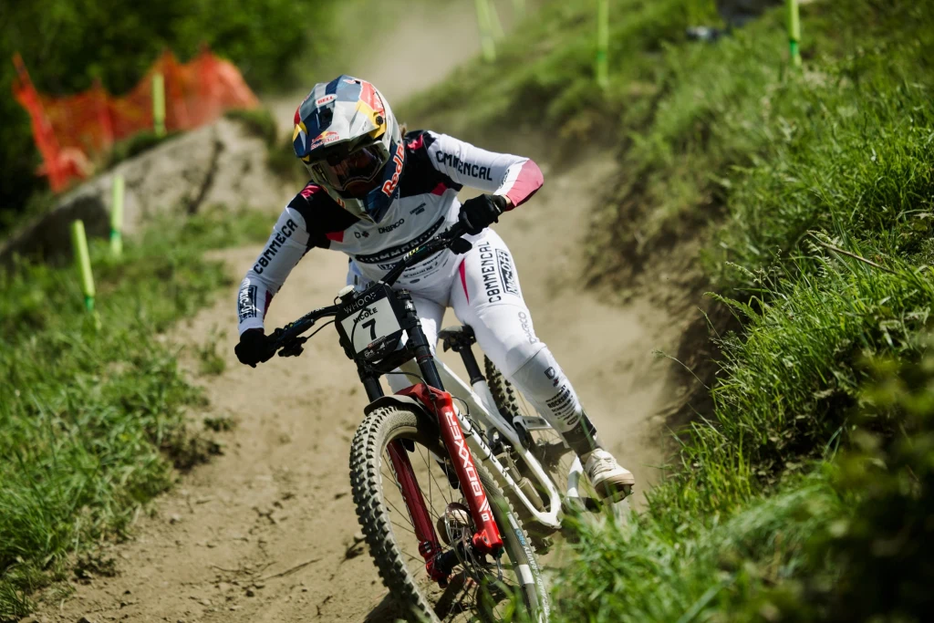 Zjazdowiec w profesjonalnym stroju i kasku pędzi rowerem górskim po zakurzonej, krętej trasie w terenie otoczonym zielenią i niskimi barierkami ochronnymi.