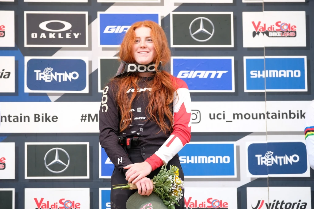 Kobieta z długimi rudymi włosami ubrana w sportowy strój rowerowy oraz ochraniacz na szyję, trzyma kwiaty i medal, stoi na podium przed ścianą z logo sponsorów podczas zawodów rowerowych.