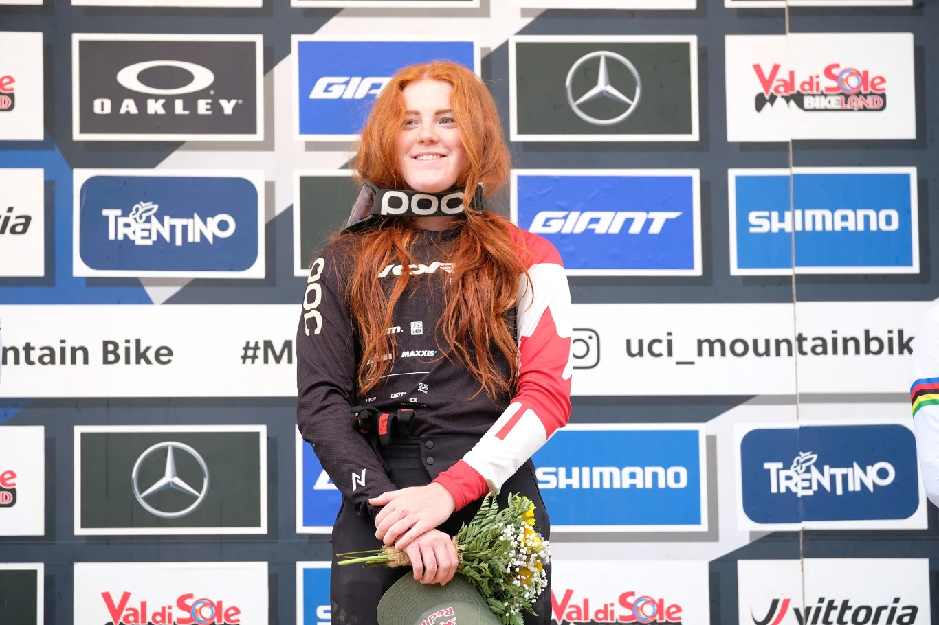 Gracey Hemstreet Kobieta z długimi rudymi włosami ubrana w sportowy strój rowerowy oraz ochraniacz na szyję, trzyma kwiaty i medal, stoi na podium przed ścianą z logo sponsorów podczas zawodów rowerowych.