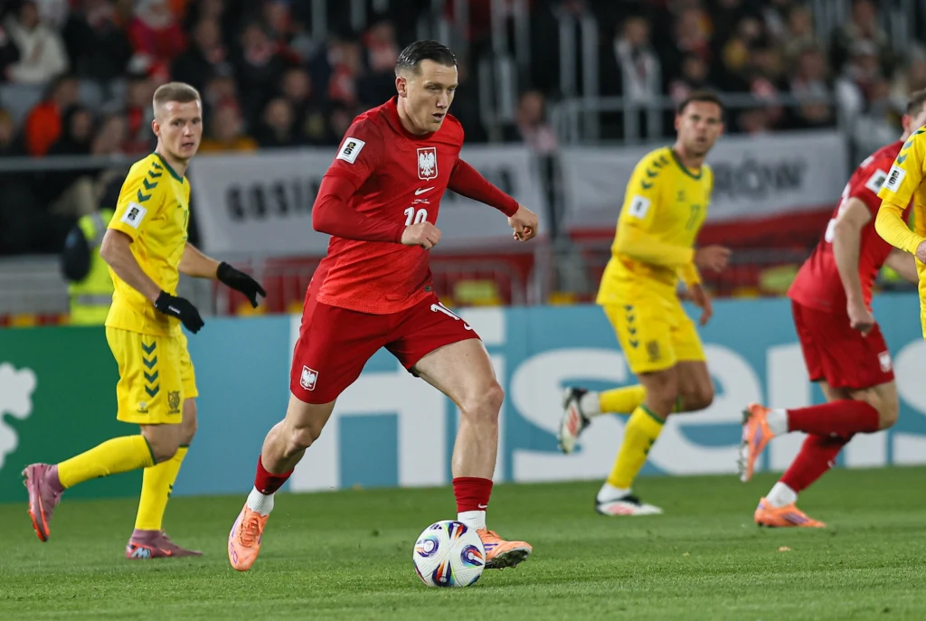 Litwa - Polska. Na zdjęciu Piotr Zieliński Piłkarz w czerwonym stroju reprezentacji Polski drybluje z piłką na boisku podczas meczu, otoczony przez zawodników przeciwnej drużyny w żółtych strojach, trybuny w tle wypełnione kibicami.