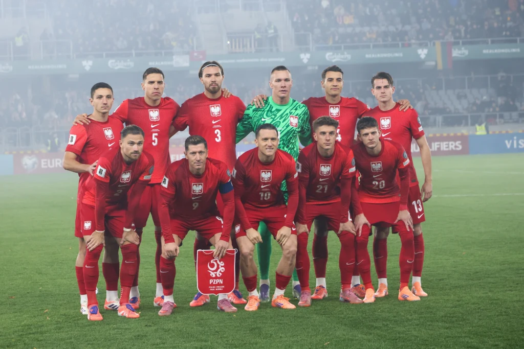 Polska reprezentacja w piłce nożnej pozuje w kompletnych czerwonych strojach na murawie stadionu tuż przed rozpoczęciem meczu, zawodnicy ustawieni w dwóch rzędach, w tle widać trybuny pełne kibiców oraz rozmytą flagę Niemiec.