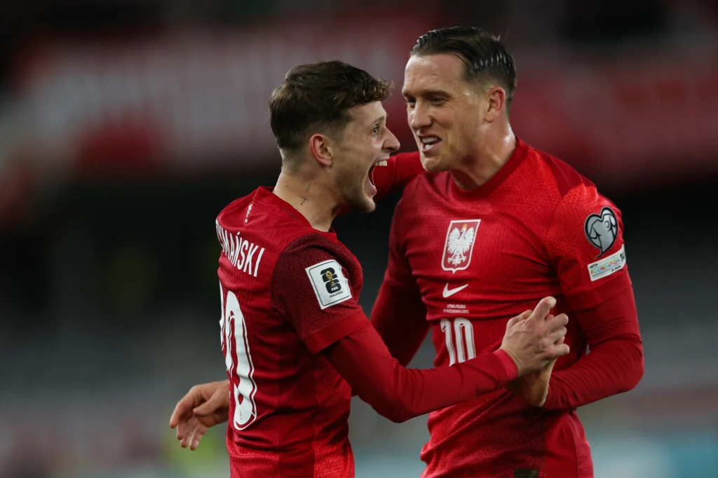 Dwóch piłkarzy reprezentacji Polski w czerwonych strojach świętuje zdobycie bramki na stadionie, wyrażając radość oraz emocje związane z meczem piłki nożnej. Jeden zawodnik z numerem 20 na koszulce, drugi z numerem 10.