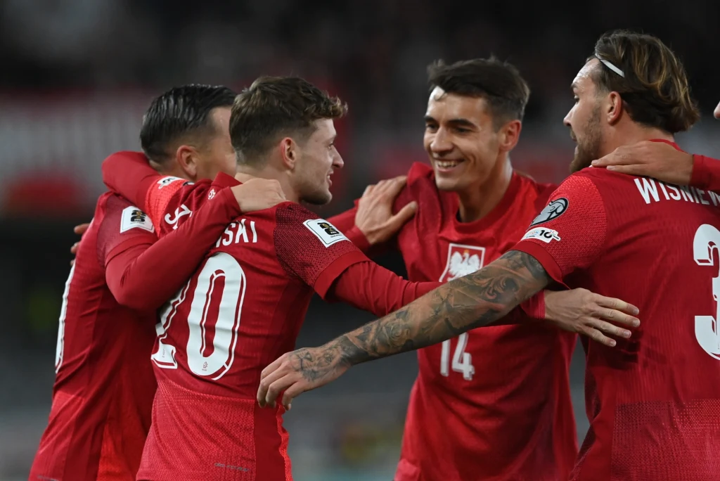 Już po kwadransie było radośnie. Sebastian Szymański (nr 10) trafił do siatki bezpośrednio z kornera Grupa polskich piłkarzy świętuje wspólnie sukces na boisku, uśmiechnięci zawodnicy przytulają się, ubrani w czerwone stroje reprezentacji narodowej.