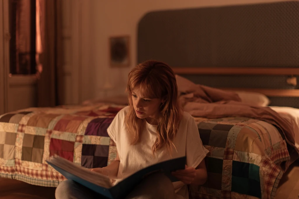 Młoda kobieta siedzi na podłodze w przytulnym, ciepło oświetlonym pokoju i skupiona przegląda duży, niebieski album. W tle widoczny jest łóżko nakryte kolorową narzutą patchworkową oraz poduszki.