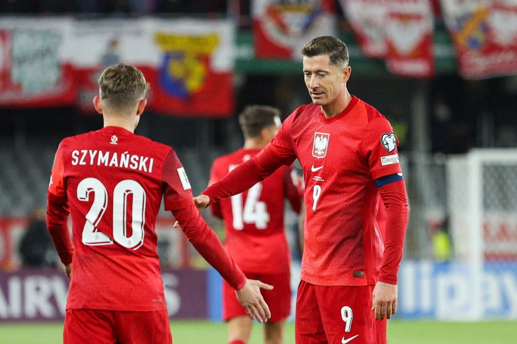 reprezentacja Polski Dwóch piłkarzy reprezentacji Polski w czerwonych strojach, jeden z nich ma opaskę kapitana, na stadionie widoczne są flagi i transparenty w tle.