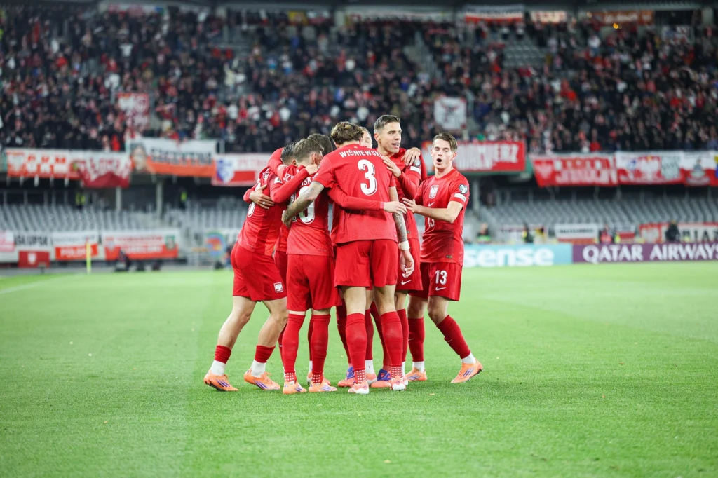 reprezentacja Polski Grupa piłkarzy ubranych w czerwone stroje świętuje na murawie stadionu po zdobyciu bramki, otoczona przez kibiców na trybunach z biało-czerwonymi flagami.