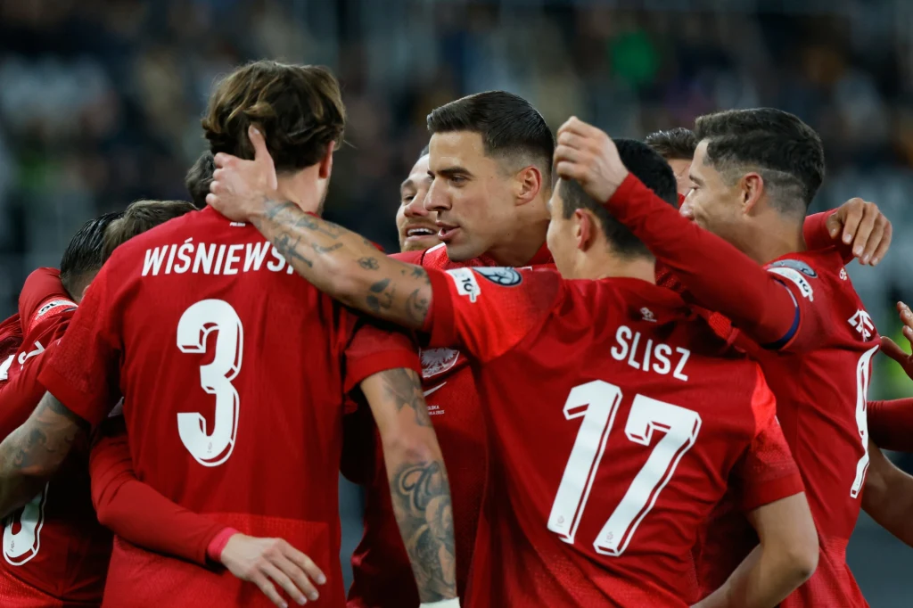 Grupa piłkarzy w czerwonych koszulkach świętuje razem na boisku, obejmując się po zdobyciu gola, widoczne nazwiska i numery na plecach zawodników.