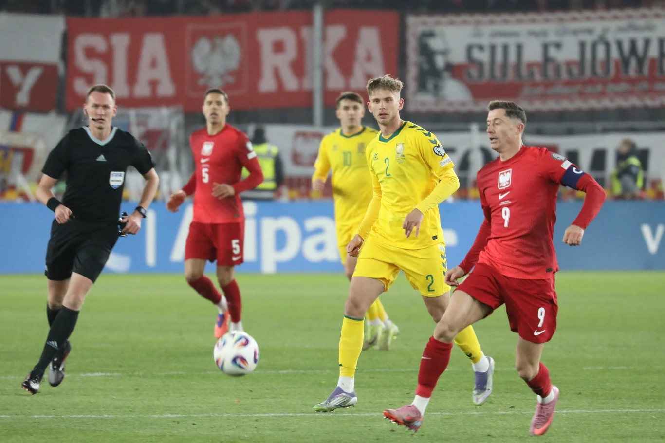 Reprezentacja Polski w meczu z Litwą (12.10.2025) Zawodnicy drużyn w żółtych i czerwonych strojach rywalizują na boisku piłkarskim podczas meczu międzynarodowego, na pierwszym planie piłkarz z numerem 9 wykonuje ruch w kierunku piłki, obok znajduje się sędzia oraz pozostali gracze obserwujący akcję.