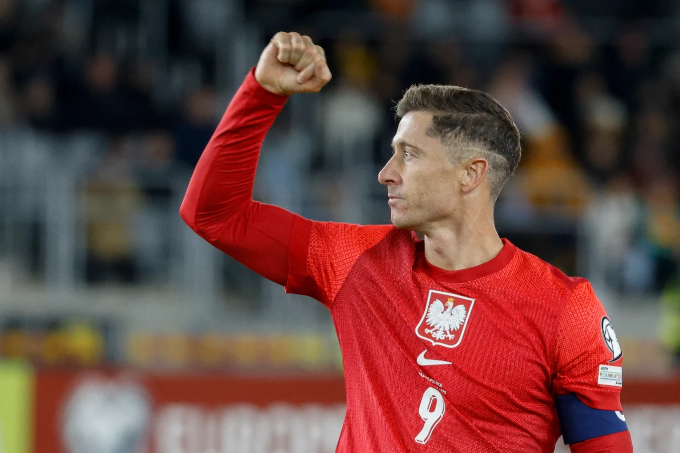 Robert Lewandowski Piłkarz w czerwonej koszulce z orłem na piersi i numerem 9 świętuje sukces unosząc zaciśniętą pięść w górę, na ramieniu opaska kapitana, w tle rozmazany stadion.
