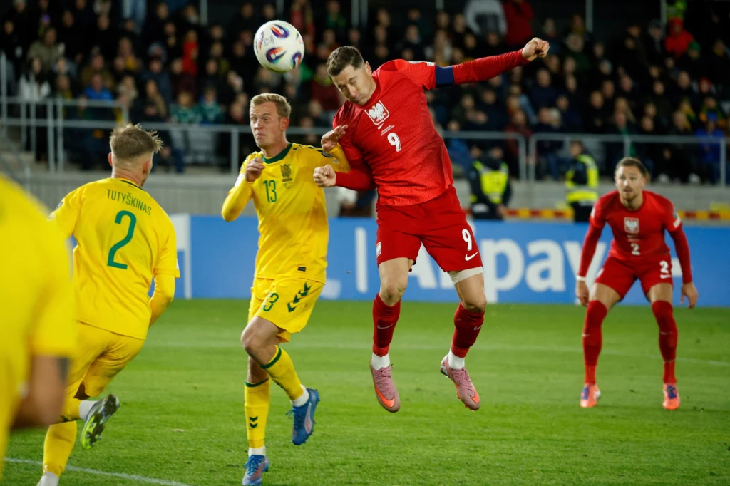 Robert Lewandowski strzelający bramkę Litwie (12.10.2025) Piłkarz w czerwonym stroju oddaje strzał głową podczas rywalizacji z zawodnikami w żółtych strojach na tle trybun i murawy stadionu, w tle widoczni pozostali gracze oraz kibice.