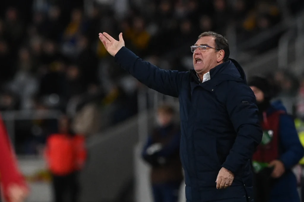 Jan Urban, w roli selekcjonera wciąż niepokonany Mężczyzna w ciemnej kurtce energicznie gestykuluje, stojąc na tle rozmytej publiczności na stadionie sportowym.