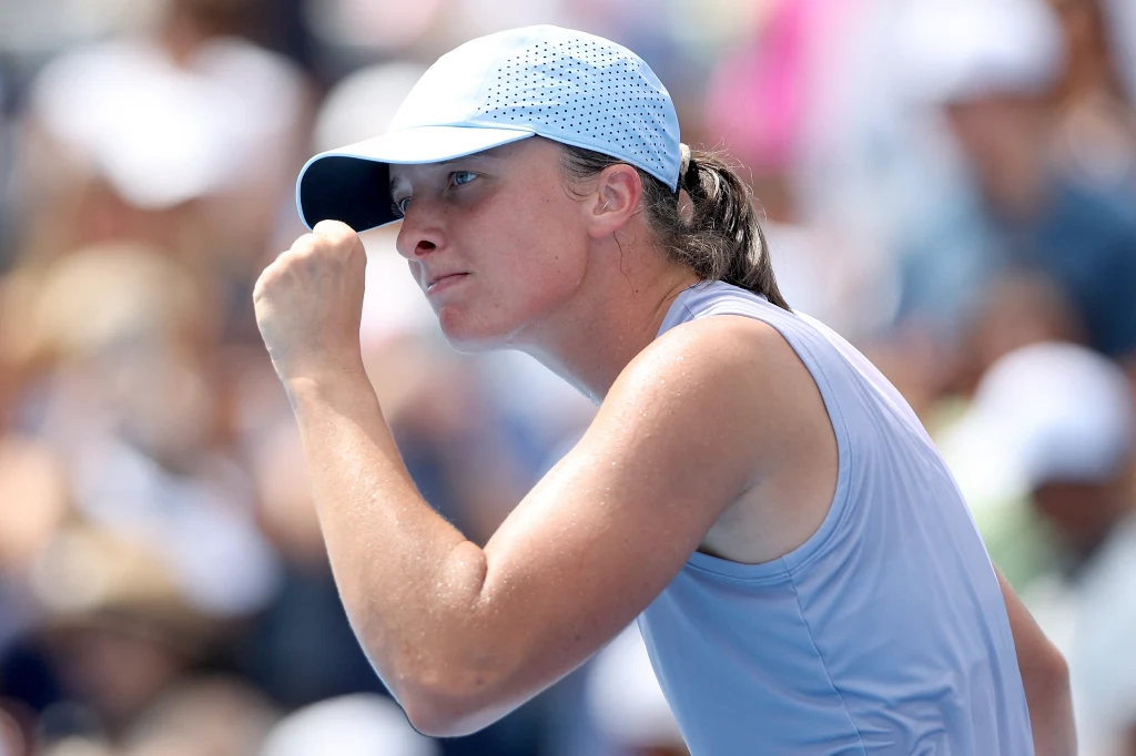 Młoda kobieta w sportowym stroju i jasnej czapce wykonuje gest zaciśniętej pięści, wyrażający determinację i triumf, na tle rozmytego tłumu kibiców.