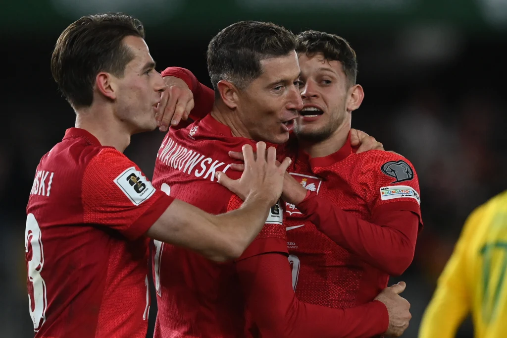 Jakub Kamiński, Robert Lewandowski i Sebastian Szymański podczas meczu Litwa - Polska Trzech piłkarzy w czerwonych koszulkach reprezentacji Polski świętuje zdobycie bramki, obejmując się i wyrażając radość, na drugim planie widać fragment zawodnika w żółtej koszulce przeciwnika.