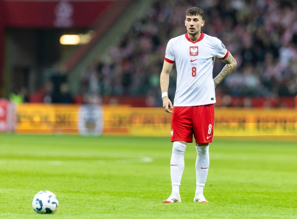 Jakub Moder Piłkarz reprezentacji Polski w biało-czerwonym stroju stoi na murawie stadionu w trakcie meczu, obok niego leży piłka, w tle widać trybuny pełne kibiców.