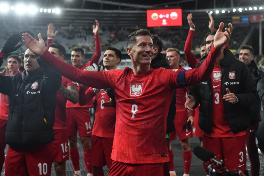 Reprezentanci Polski na czele z Robertem Lewandowskim Piłkarze reprezentacji Polski w czerwonych strojach świętują zwycięstwo na stadionie, na czele z kapitanem, unoszącym ręce w geście triumfu, wokół nich widownia i światła stadionu