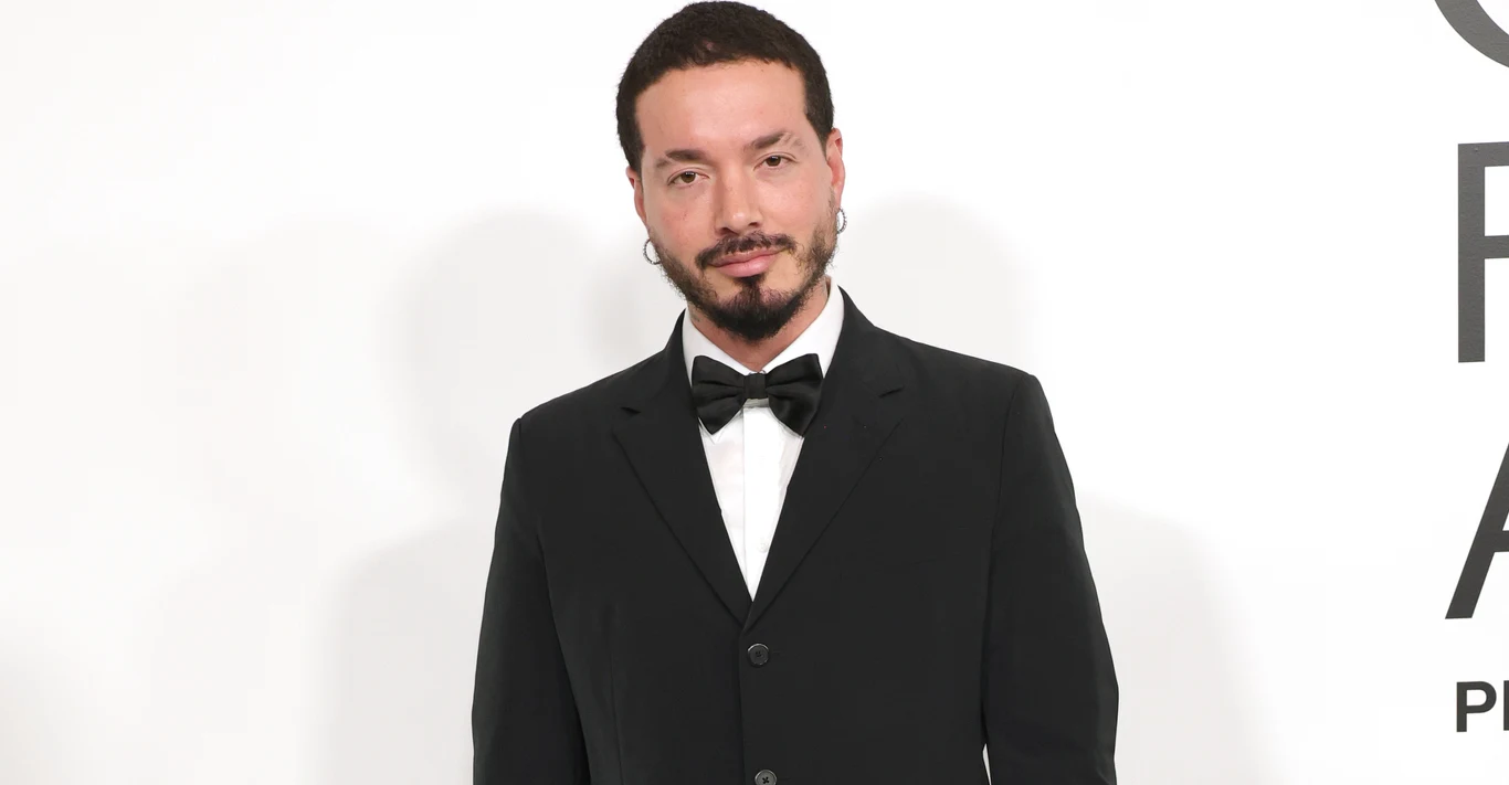 J Balvin Mężczyzna w eleganckim, czarnym garniturze z muchą, stojący na jasnym tle, z lekkim uśmiechem na twarzy, widoczny portretowo od pasa w górę.