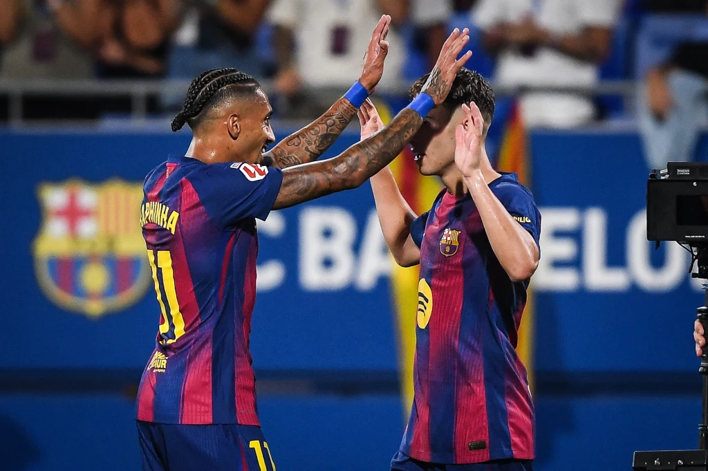 Raphinha i Fermin Lopez Dwóch piłkarzy w granatowo-bordowych strojach Barcelony świętuje razem, przybijając sobie piątki, na tle klubowego baneru