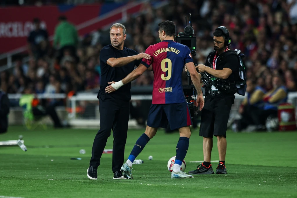 Hansi Flick i Robert Lewandowski Zawodnik drużyny piłkarskiej w stroju meczu schodzi z boiska, podając rękę trenerowi, podczas gdy w tle znajduje się operator kamery oraz publiczność na trybunach stadionu