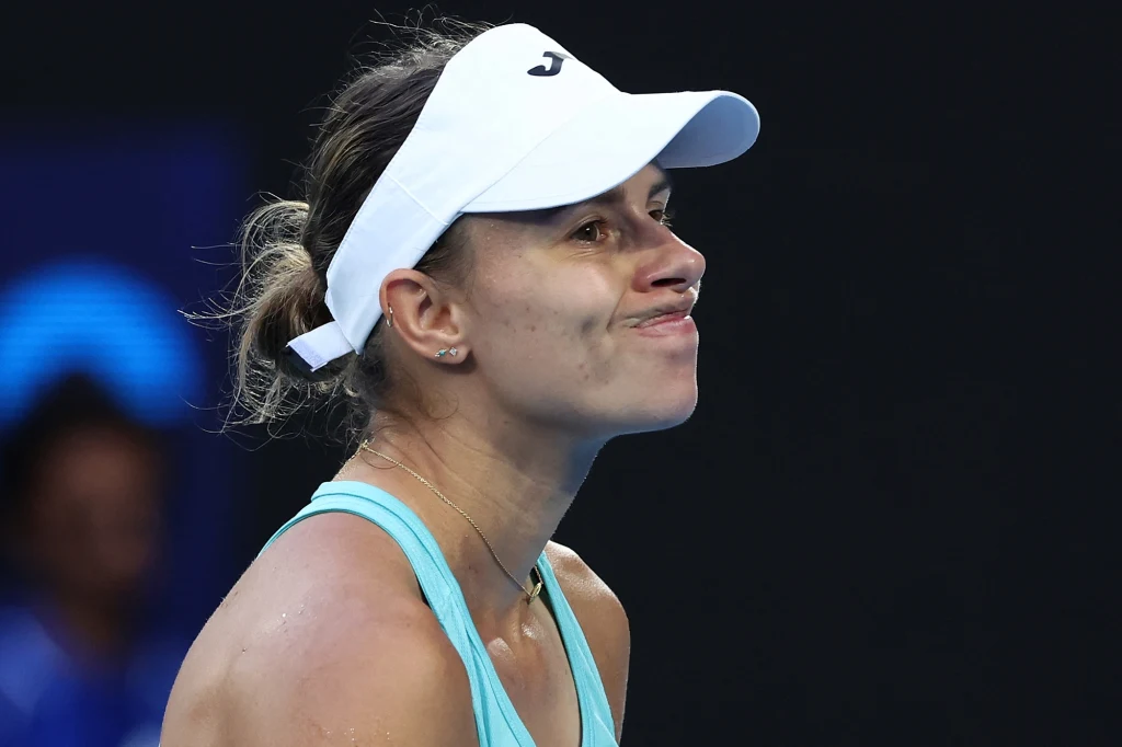 Zbliżenie na kobietę w sportowym stroju i białej czapce z daszkiem, która z wyraźną miną niezadowolenia stoi na tle ciemnego boiska tenisowego.