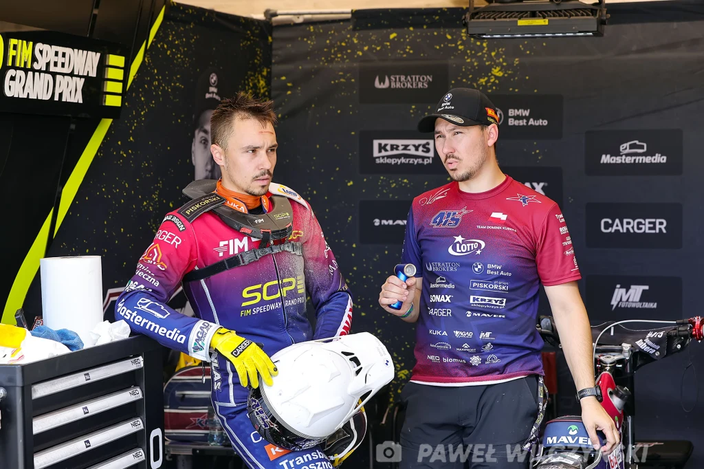 Dwóch mężczyzn ubranych w stroje żużlowe rozmawia ze sobą w boksie, jeden z nich trzyma biały kask, w tle tablice z nazwami sponsorów oraz sprzęt sportowy.