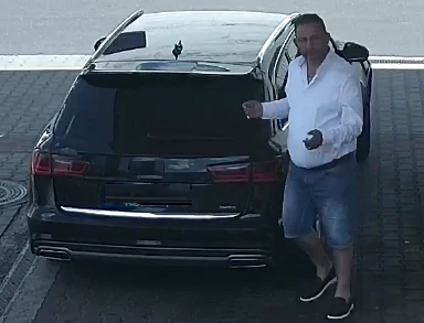 Mężczyzna w białej koszuli i dżinsowych spodenkach stoi obok ciemnego samochodu osobowego zaparkowanego na chodniku, trzyma w ręku kluczyki samochodowe.