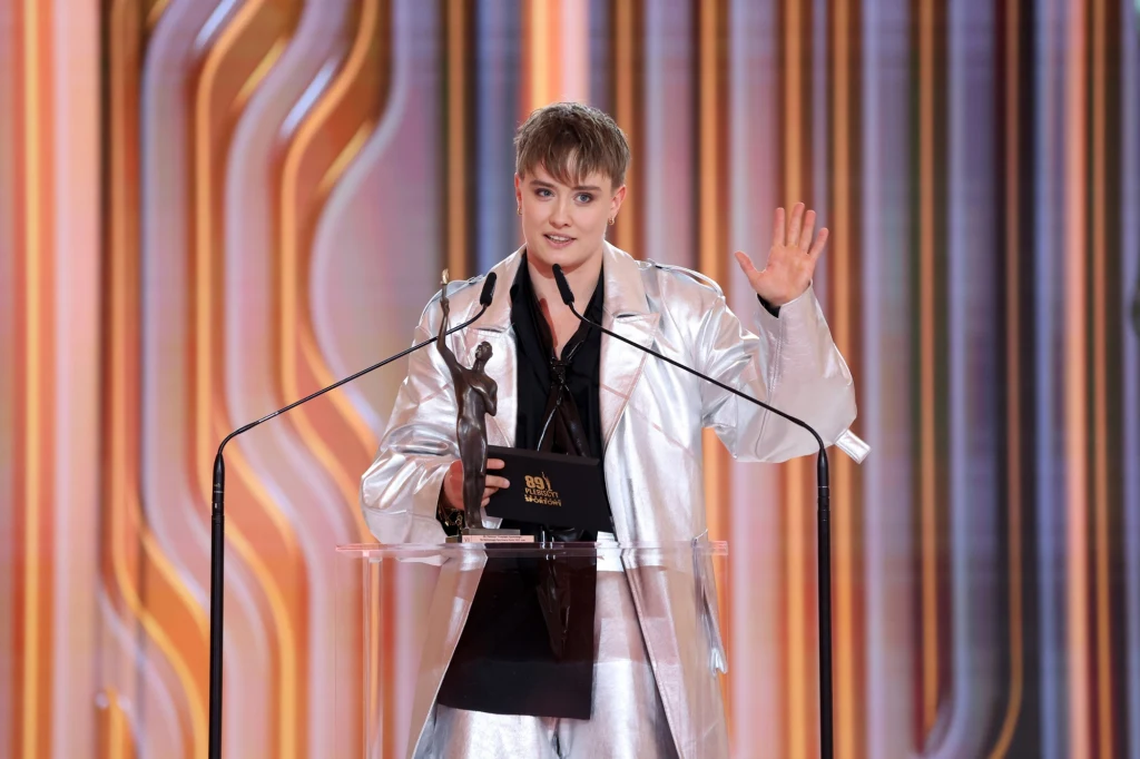 Osoba w srebrnym garniturze stoi na scenie z mikrofonem, trzyma statuetkę oraz czarną teczkę, gestykulując jedną ręką w górę na tle kolorowych, pionowych dekoracji.