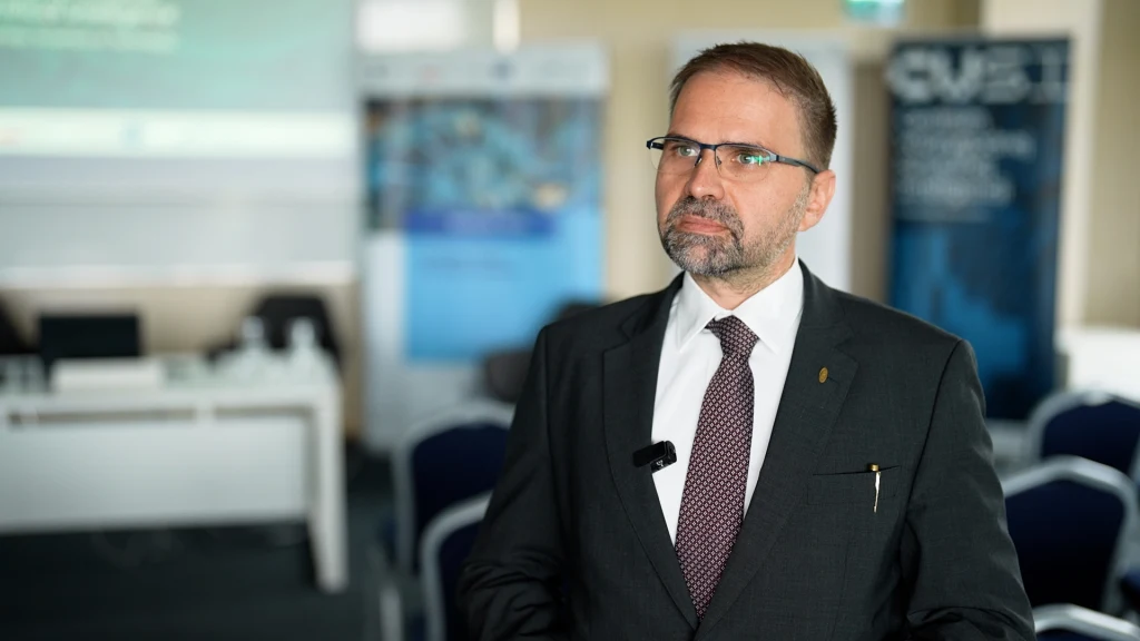 Mężczyzna w garniturze i okularach, z mikrofonem przypiętym do klapy marynarki, znajduje się w jasnym pomieszczeniu konferencyjnym z plakatami i ekranem w tle