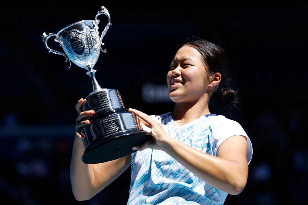 Młoda kobieta w sportowym stroju trzyma duży srebrny puchar z uśmiechem na twarzy, będąc na zewnątrz w jasnym świetle dziennym.