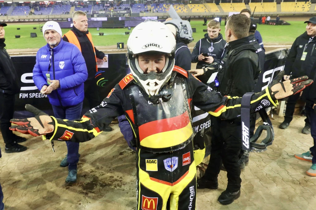 Zawodnik żużlowy w pełnym stroju i kasku gestykuluje szeroko rozłożonymi rękami po wyścigu na stadionie, w tle widoczni inni członkowie ekipy oraz trybuny z kibicami.
