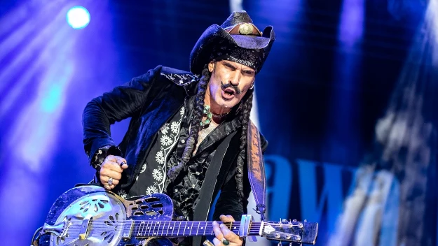 Mężczyzna w kowbojskim kapeluszu gra na metalicznej gitarze na scenie koncertowej, otoczony barwnym światłem i scenografią, wyrażając intensywne emocje mimiką twarzy.