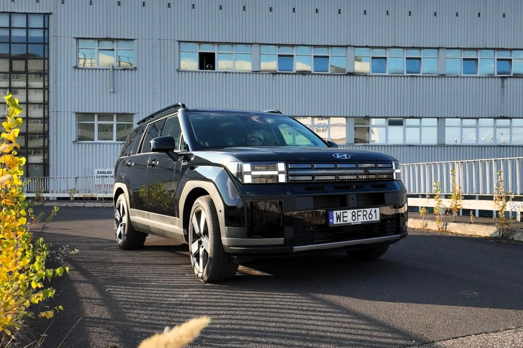 Czarny samochód Hyundai stoi zaparkowany na asfalcie przed przemysłowym budynkiem z licznymi oknami, w tle widoczne są metalowe barierki oraz fragmenty roślinności.