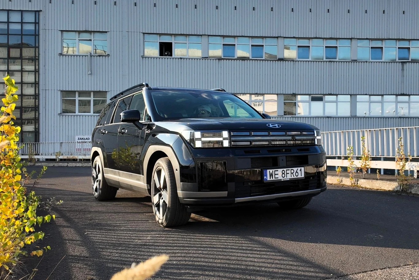 Czarny samochód Hyundai stoi zaparkowany na asfalcie przed przemysłowym budynkiem z licznymi oknami, w tle widoczne są metalowe barierki oraz fragmenty roślinności.