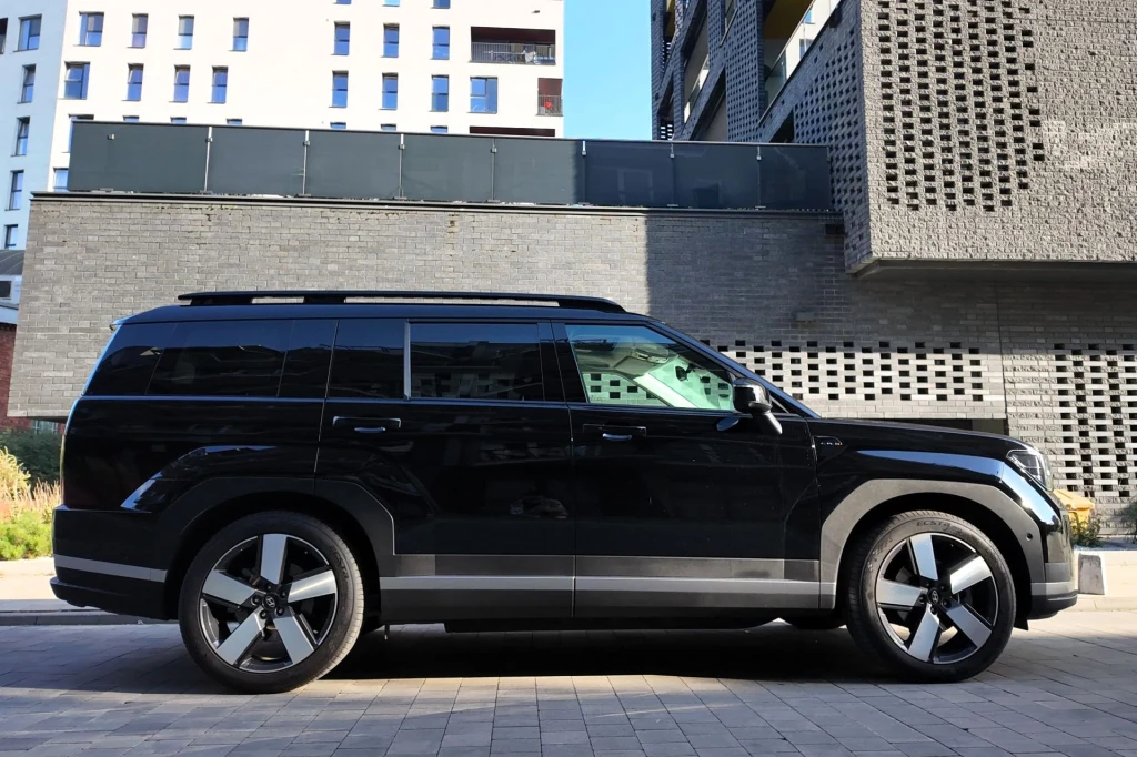 Czarny samochód SUV zaparkowany na tle nowoczesnych budynków o surowych elewacjach, w słoneczny dzień, samochód dominuje na pierwszym planie, akcentując nowoczesny design i dynamiczne linie pojazdu.