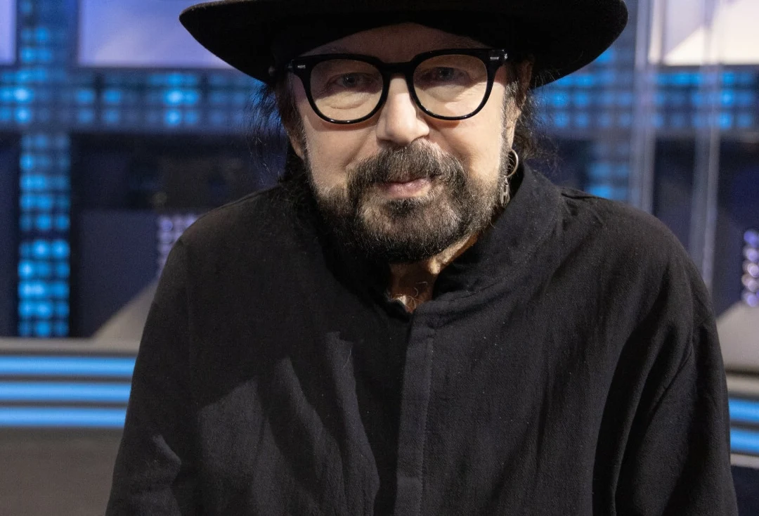 Zbigniew Hołdys zmaga się z chorobą Mężczyzna w czarnym kapeluszu, czarnej koszuli i okularach, ma brodę i wąsy, patrzy prosto w obiektyw, w tle widoczne rozmyte elementy scenografii oświetlone niebieskimi światłami.