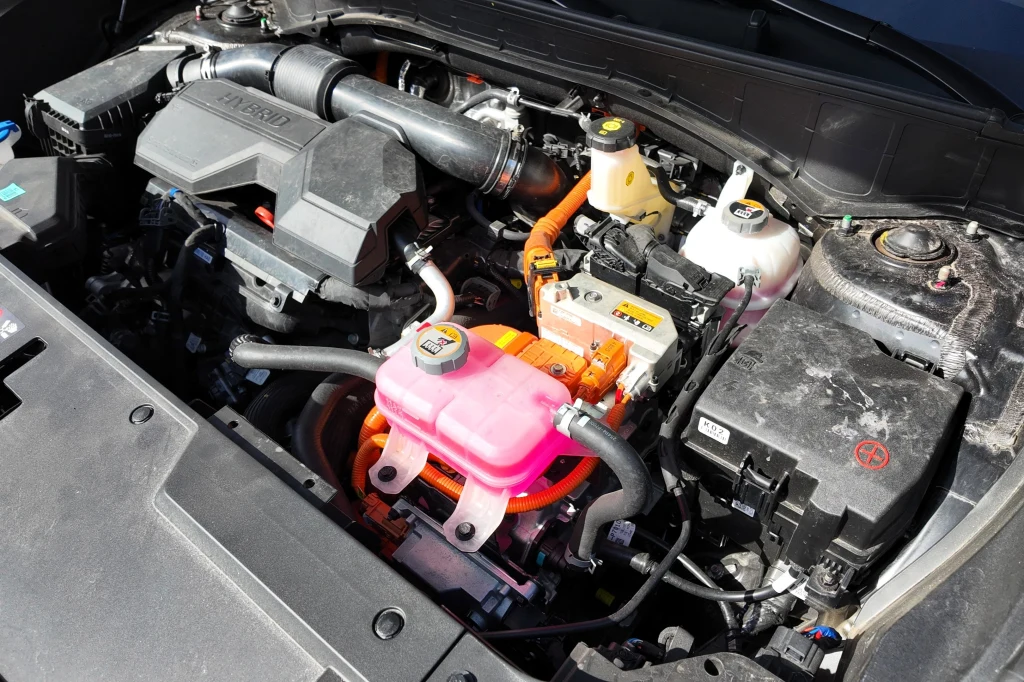 Komora silnika hybrydowego samochodu z widocznymi przewodami wysokiego napięcia, zbiornikiem płynu chłodniczego z jaskrawo różowym płynem, elementami instalacji elektrycznej oraz filtrem powietrza.