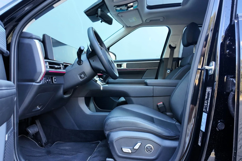 Luksusowe wnętrze samochodu z nowoczesnym panelem sterowania, czarnymi skórzanymi fotelami oraz dużymi ekranami na desce rozdzielczej, widok z otwartych drzwi kierowcy