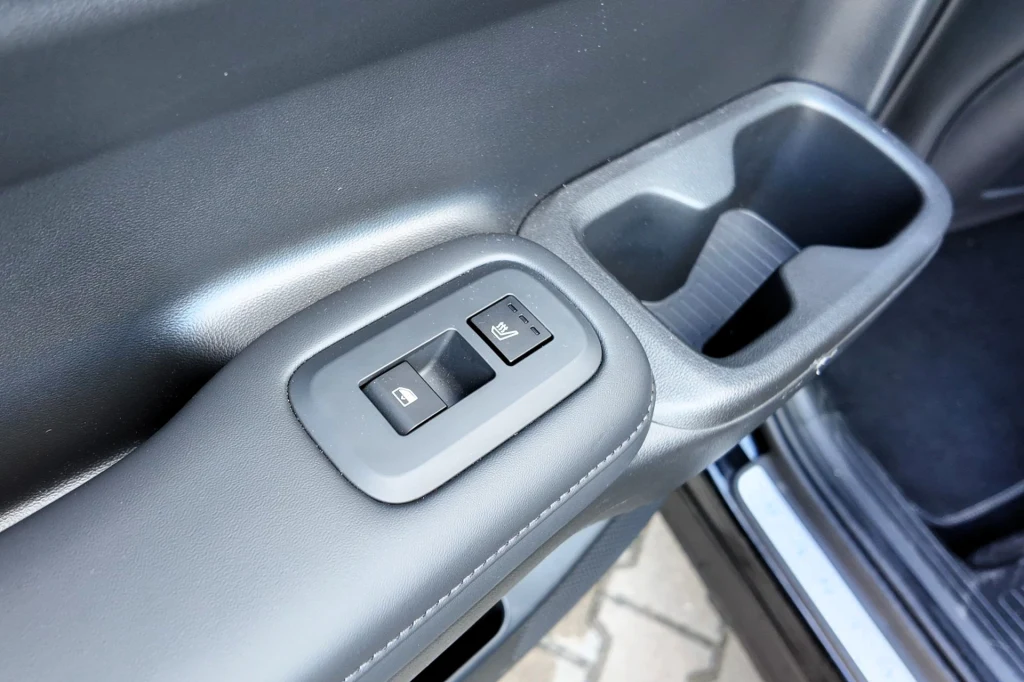 Panel sterowania szybami elektrycznymi oraz przycisk blokady szyb w samochodzie, wykonany z czarnego tworzywa z wyraźnymi oznaczeniami piktogramów, umieszczony w drzwiach pojazdu, obok schowka na drobne przedmioty.