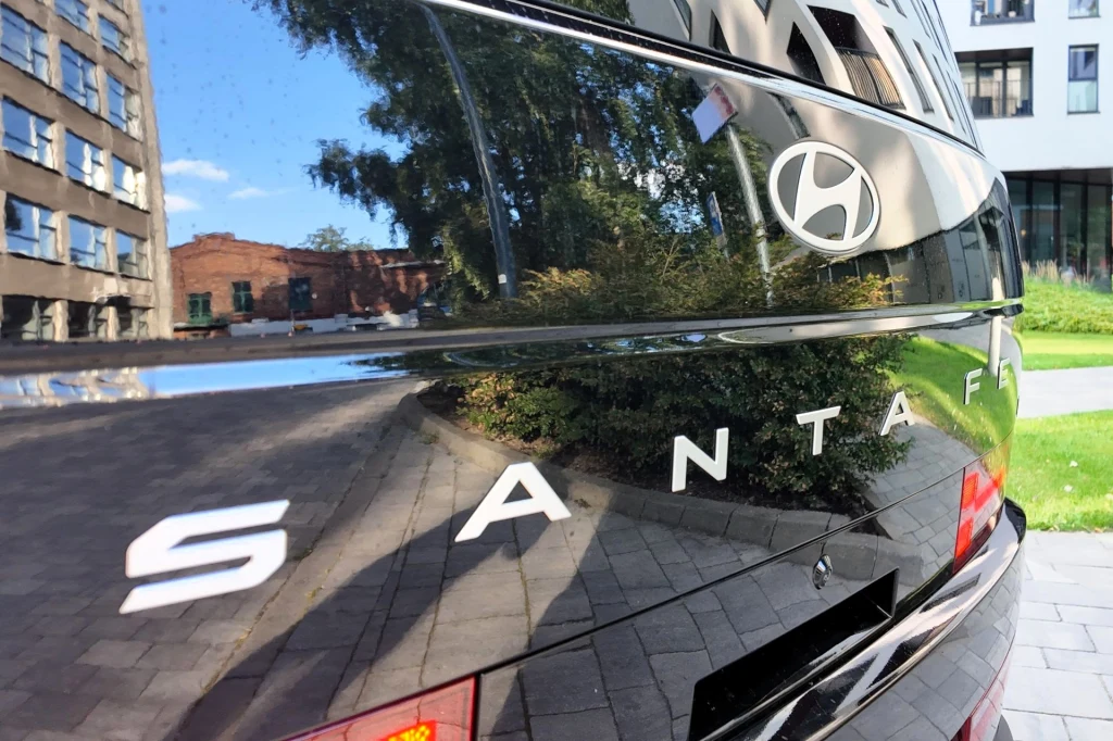 Czarny lakierowany tył samochodu Hyundai Santa Fe z wyraźnie widocznym logo oraz odbiciem otoczenia, w tle nowoczesne budynki i zieleń.