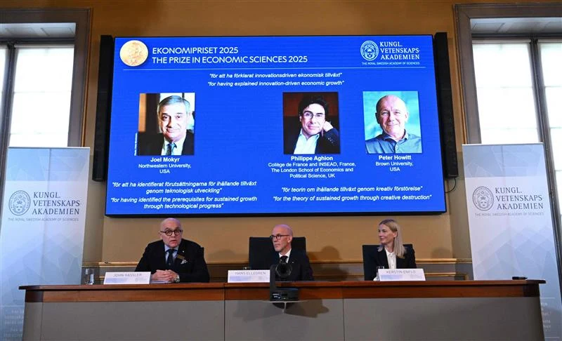 Trzech członków komitetu noblowskiego siedzi przy stole konferencyjnym z mikrofonami i tabliczkami z nazwiskami, za nimi duży ekran z informacją o laureatach Nagrody Nobla w dziedzinie ekonomii na tle niebieskiego tła oraz zdjęcia trzech nagrodzonych o...